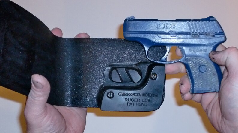 May include: A black leather holster with a blue Ruger LC9 pistol inside. The holster has the text "KEVINSCONCEALMENT.COM" and "RUGER LC9 PAT PEND" on it.