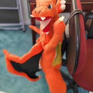 May include: Orange and black plush toy of a dragon with a wide open mouth and sharp teeth. The toy is hanging from a chair with a black strap.