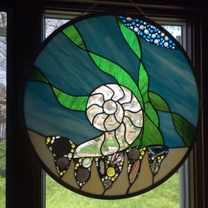 Puede incluir: Una ventana redonda de vitrales que representa una escena submarina con una concha de nautilus, algas verdes y burbujas azules. La ventana está enmarcada en metal negro.