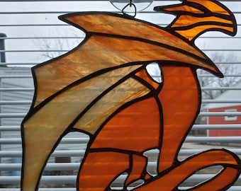 May include: A stained glass dragon with orange and yellow wings and a red body. The dragon is hanging from a window with a suction cup.
