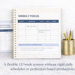 May include: A spiral-bound planner with a weekly focus layout, featuring sections for daily tasks and self-talk affirmations. Includes a separate page for law school applications. Gold pencils and a pen holder are also visible.