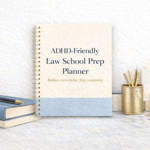 May include: An ADHD-Friendly Law School Prep Planner with a light blue and cream cover, spiral binding, and gold accents. The planner is next to a stack of blue books, a pen, a gold pencil holder with pencils, and paperclips.