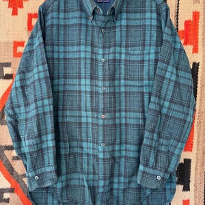 May include: A teal and black plaid button-down shirt. The long-sleeved shirt has a classic collar and buttoned cuffs. The shirt is made of a soft, woven fabric. The Pendleton label is visible on the collar.