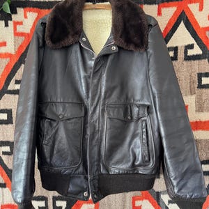 Puede incluir: Una chaqueta bomber de cuero negro vintage con cuello de piel marrón y forro interior color crema. La chaqueta tiene dos bolsillos delanteros, cierre de cremallera y dobladillo y puños de canalé. La chaqueta está colgada en una percha de madera.