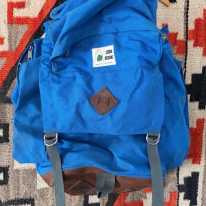 May include: A blue backpack with a brown leather bottom and accents. The backpack has gray straps and a flap with the text "Sierra Designs Berkeley, California". The backpack is hanging on a wooden hanger.