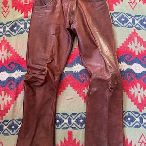 May include: Brown leather pants with a classic five-pocket design. The pants have a button and zipper closure and show signs of wear. The leather has a rich, warm tone, and the pants are laid flat on a patterned textile.