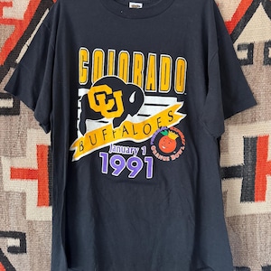 May include: Black t-shirt featuring the text "COLORADO BUFFALOES" in yellow and white, with a buffalo logo. The shirt also includes "January 1 1991" and an "Orange Bowl" graphic.