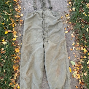 May include: Olive green vintage work overalls with suspenders, two front pockets, and button closures. The overalls have a faded appearance and show signs of wear. The letters "U.S.N." are visible on the upper right chest area.