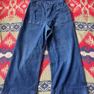 May include: Vintage blue denim pants with wide legs. The pants have two large front pockets and a button closure. The denim has a faded appearance, suggesting wear. The pants are laid flat on a patterned textile.