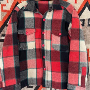 Könnte beinhalten: Vintage karierte Jacke in Rot, Schwarz, Creme und Grün. Die Jacke hat einen Kragen, zwei Brusttaschen und rote Knöpfe. Die Jacke hängt an einem Kleiderbügel.