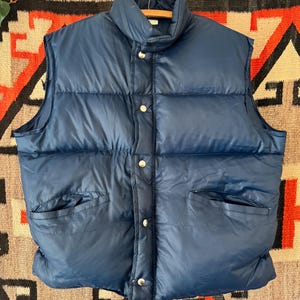May include: A navy blue puffer vest with a high collar and two front pockets. The vest has a quilted design and is fastened with silver snap closures. It is hanging on a wooden hanger.