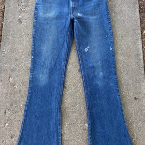 May include: A pair of blue denim jeans featuring a faded wash, a button fly closure, and a flared leg silhouette. The fabric displays light whiskering at the hips and subtle distressed wear marks on the upper thigh area, with a classic five-pocket design.
