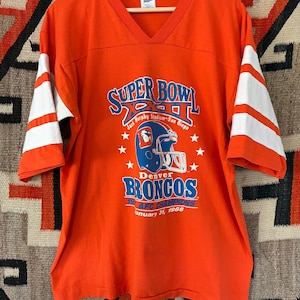May include: An orange V-neck t-shirt with white stripes on the sleeves. The front features a graphic with the text Super Bowl XXII, Jack Murphy Stadium-San Diego, Denver Broncos, 1987 AFC Champions, and the date January 31, 1988, with a helmet.