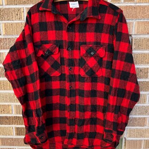 Könnte beinhalten: Rotes und schwarzes kariertes Flanellhemd mit Knopfleiste und zwei Brusttaschen. Das Hemd trägt ein Etikett mit der Aufschrift "Fansett Glove Works, Inc. & More Cotton".