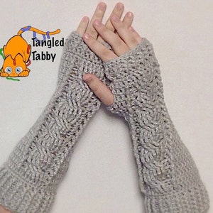 Könnte beinhalten: Ein Paar graue Fingerlose Handschuhe mit einem Zopfmuster. Die Handschuhe sind aus weichem Garn gefertigt und haben ein warmes, gemütliches Gefühl.
