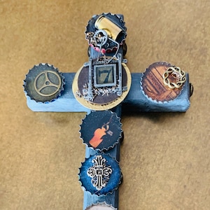 May include: A wooden cross decorated with bottle caps. The caps feature various designs, including a dog, a cross, a number seven, and a gear. The cross is painted a dark blue color.