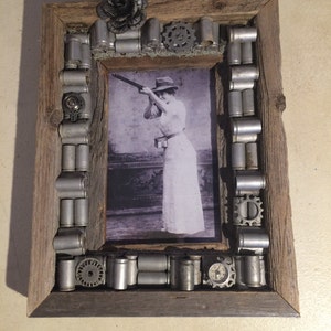 May include: A rustic wooden picture frame decorated with metal casings, gears, and a metal flower. The frame contains a black and white photograph of a woman holding a rifle.