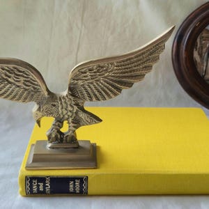May include: A brass eagle sculpture with outstretched wings sits atop a bright yellow book. The book has a dark blue spine with the text "DANCE and SKYLARK" and "JOHN MOORE". An oval framed landscape painting is in the background.