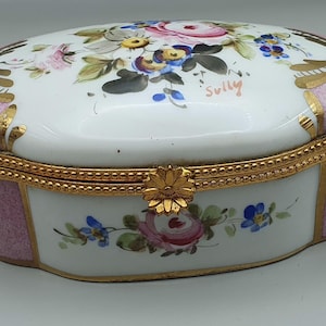 May include: Oval porcelain trinket box with a white lid and pink sides. The lid is decorated with hand-painted flowers in pink, blue, and yellow, with gold accents. The box has a gold-colored metal trim and a floral clasp. The word "Sully" is written on the lid.