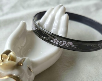 May include: A black bangle bracelet featuring a delicate, light-colored floral pattern with thin, flowing lines. The bracelet has a smooth, dark finish and is displayed on a white ceramic hand-shaped stand adorned with a gold-trimmed rose detail.
