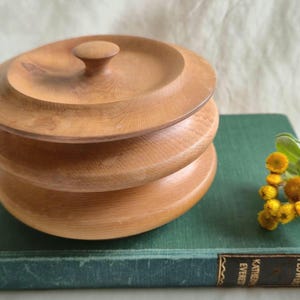 May include: A round, light brown wooden lidded container with a smooth, tiered design and a small central knob on the lid. The container features visible wood grain patterns and a polished finish, showcasing a stacked, circular construction.