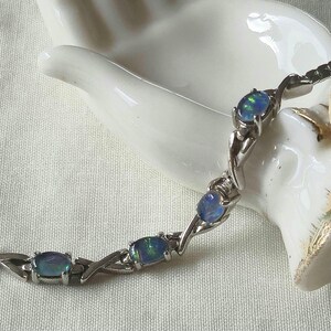 May include: A silver-toned bracelet featuring a series of oval-shaped dark blue opals with iridescent flashes of green and blue. The stones are set in metal links that alternate with polished silver-toned X-shaped connectors in a repeating pattern.