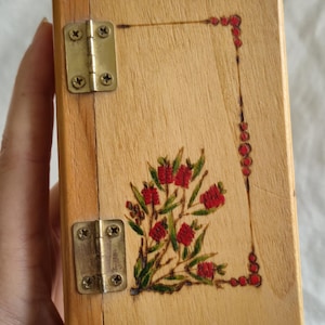 May include: A rectangular light-coloured wooden box featuring two brass-coloured metal hinges on the left side. The front surface is decorated with a wood-burned illustration of red bottlebrush flowers with green leaves and a dotted border design.