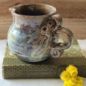 May include: A handcrafted ceramic jug with a distinctive handle. The jug displays a mix of blue, brown, and cream hues. It rests on a vintage book with a green cover. A small bunch of yellow flowers is in the foreground.