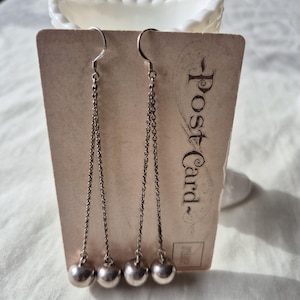 May include: Silver dangle earrings with ball charms. The earrings hang from a chain and are displayed on a vintage postcard. The postcard has the word "Postcard" printed on it.