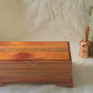 May include: A wooden box with a hinged lid, showing a natural wood grain. A copper bell, a vintage book, and a small potted plant are arranged nearby. The box is rectangular and rests on small feet.