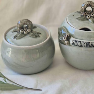 May include: Two round, light green ceramic containers with lids. The larger container features the word SUGAR embossed on the side. Both lids are decorated with sculpted green leaves and small white berries. A sprig of green leaves lies nearby.