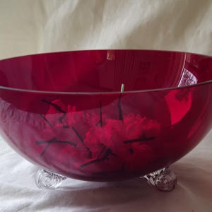 May include: A large, deep red glass bowl with a rounded shape, supported by three clear, decorative feet. The bowl contains red flowers and stems. The bowl is likely for decorative use.