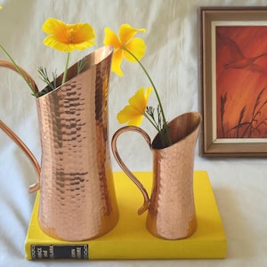 May include: Two copper pitchers with a hammered texture, one large and one small, each with a curved handle. Yellow flowers are arranged in the pitchers. A framed painting with an orange and brown colour scheme is in the background.