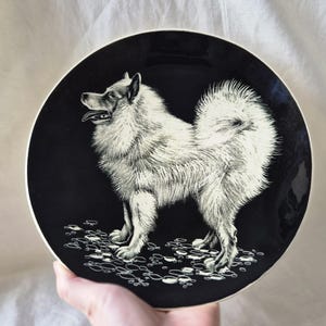 May include: A black ceramic plate with a detailed illustration of a fluffy, light-coloured dog. The dog is standing on a surface with a pattern of small, white shapes. The plate has a white rim.