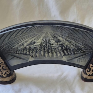 May include: An arched sculpture with a detailed grayscale depiction of a large crowd within a structure. The dark brown base features gold scrollwork and a scroll-like design on each end.