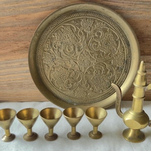 May include: A vintage brass serving set featuring a large round tray with an embossed floral design, a small pitcher, and six small martini-style glasses. The set is displayed on a white surface against a wooden background.