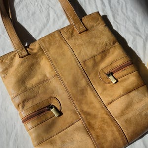 May include: A tan leather tote bag with two zipped pockets and two shoulder straps. The bag is rectangular with a vertical seam down the centre and horizontal seams across the front. The zips have gold-coloured pulls.
