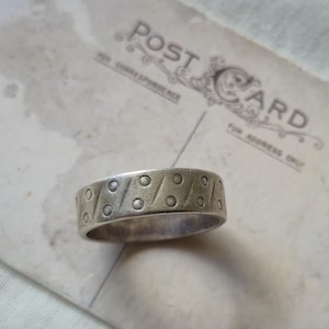 May include: A silver ring featuring a repeating pattern of circles and diagonal lines. The ring is placed on a vintage postcard with the words "POST CARD" printed on it.