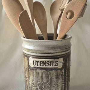 May include: A cylindrical ceramic crock with a speckled beige and brown glaze, featuring the word UTENSILS embossed on a raised rectangular plaque. The exterior has vertical textured grooves. It holds several light-coloured wooden kitchen utensils.