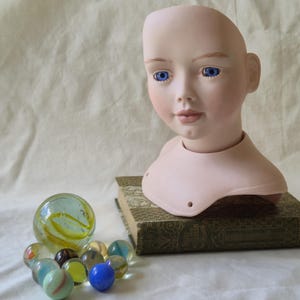May include: A porcelain doll head with blue eyes and a neutral skin tone. The doll head rests on a vintage book, with a collection of colorful marbles in the foreground. The marbles include various colors and patterns.