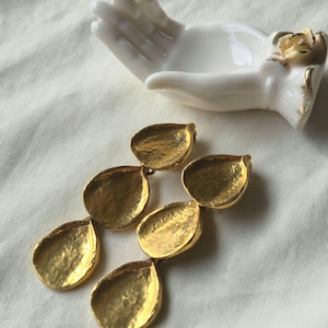 May include: Gold-coloured, teardrop-shaped earrings with a textured surface. The earrings are arranged in a cascading design. A white ceramic hand-shaped dish with gold accents is in the background.