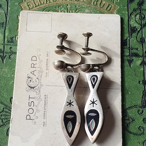 May include: Vintage silver-tone clip-on earrings. Each earring has a fish shape with black enamel and turquoise accents. The earrings are on a vintage postcard with the text "POST CARD" and "ELLAS ROSE BUD".