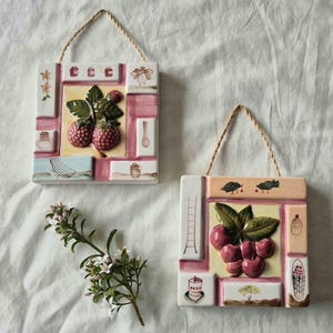 May include: Two square ceramic wall plaques with rope hangers. One features a raised raspberry motif with painted icons of a cake, chair, and spoon. The other displays a raised cherry motif with painted icons of a ladder, cupcake, and basket.