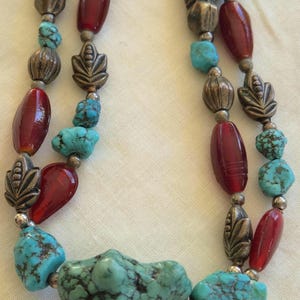 May include: A multi-strand necklace featuring irregular turquoise stones with dark matrix, smooth deep red oval beads, and antiqued silver-toned metal beads shaped like leaves and textured spheres, arranged in a repeating pattern along the strands.