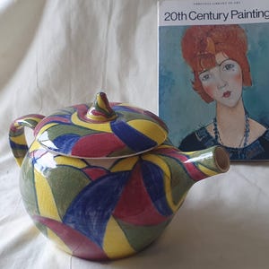 May include: A ceramic teapot featuring a vibrant, multi-coloured abstract pattern of yellow, blue, green, and red shapes. The teapot has a matching lid with a pointed handle and a curved spout. Beside it sits a book titled 20th Century Painting.