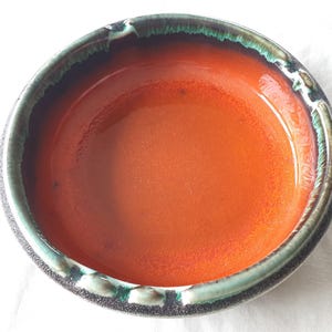 May include: A round ceramic bowl featuring a vibrant orange interior glaze. The rim is decorated with a dark brown and teal drip pattern that extends slightly over the edge. The exterior surface has a textured, dark brown finish with visible speckles.