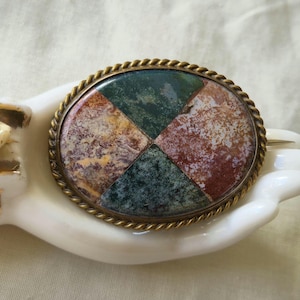 May include: Oval brooch with a mosaic design in shades of green, red, and tan, set in a gold-toned metal frame with a rope-like border. The brooch is displayed on a white ceramic hand with a gold rose detail.