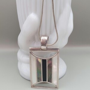 May include: A silver pendant necklace. The rectangular pendant has a geometric design with white, black, and iridescent mother-of-pearl inlays. The necklace features a fine silver chain.