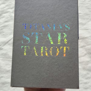 May include: A gray book titled "TITANIA'S STAR TAROT" with holographic lettering. The book is held by a person wearing a gold bracelet. The book's cover is a solid gray color.