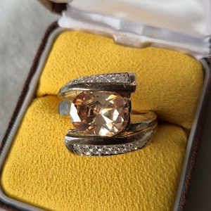 May include: A silver-toned ring featuring a large, oval-cut peach-coloured gemstone at the centre. The band is split into two curved sections, each accented with a row of small, clear, sparkling stones along the outer edges of the metal setting.